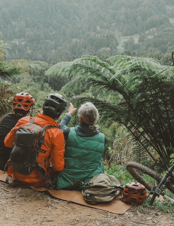 Three people sitting on a log in a forest, looking at a mountain view with bicycles nearby.